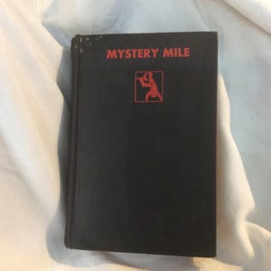May include: A black hardcover book with the title "MYSTERY MILE" printed in red on the front cover. A red illustration of a person holding a magnifying glass is also on the cover.
