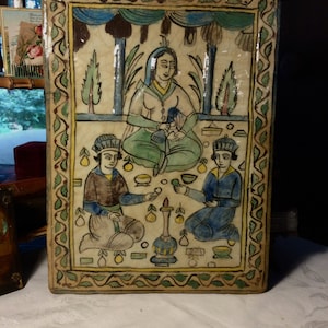 May include: Rectangular ceramic tile with a hand-painted illustration. The artwork depicts a seated figure with two attendants, all in traditional attire. The color palette includes blues, greens, and yellows, with a decorative border.