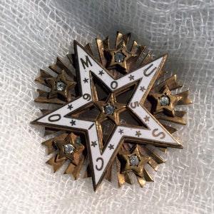 May include: A gold-toned star-shaped pin with a white enamel center. The center features a five-pointed star with the letters "M.O.O.S.U.C." around the perimeter. The star is surrounded by smaller stars with rhinestones.