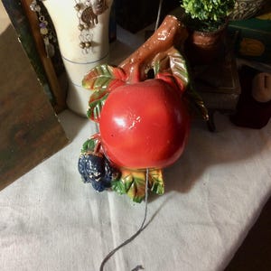 May include: A ceramic apple-shaped wall decoration. The apple is bright red with green leaves and a brown stem. A cluster of blue grapes is attached. A gray string hangs from the apple.