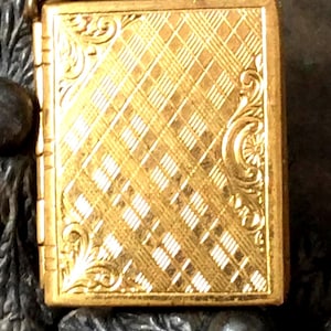 May include: A rectangular gold-toned metal locket featuring a diagonal plaid pattern across the center. The edges are decorated with embossed scrollwork and leaf motifs. The object has a visible hinge on the left side and a metallic finish.
