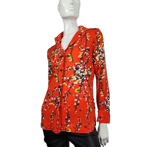 Könnte beinhalten: Ein langärmliges, geknöpftes Hemd in leuchtendem Orange, verziert mit einem Blumenmuster mit weißen, blauen, gelben und grünen Akzenten. Das Hemd hat einen klassischen Kragen und Manschetten und wird auf einer Schaufensterpuppe präsentiert.