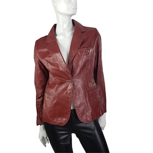 Puede incluir: Un blazer granate con cierre de un solo botón, solapas con muescas y dos bolsillos delanteros. La chaqueta se exhibe en un maniquí. El blazer tiene una apariencia texturizada similar al cuero.