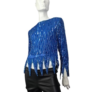 Puede incluir: Una blusa de lentejuelas azul real de manga larga con un patrón en zigzag único. La blusa presenta un cuello redondo y un dobladillo festoneado. Las lentejuelas están dispuestas en un diseño vertical y diagonal, creando un efecto brillante. La prenda se exhibe en un maniquí.