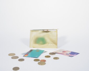 Può includere: Un portafoglio in pelle dorata con una piccola chiusura in metallo, insieme a carte di credito e monete sparse su una superficie bianca.