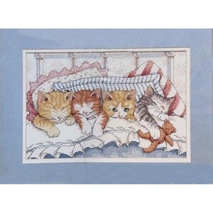 May include: A cross-stitch embroidery featuring four kittens sleeping in a bed under a striped blanket. The kittens are orange, ginger, and grey tabby, with one holding a small brown teddy bear. The artwork is framed with a light blue mat.