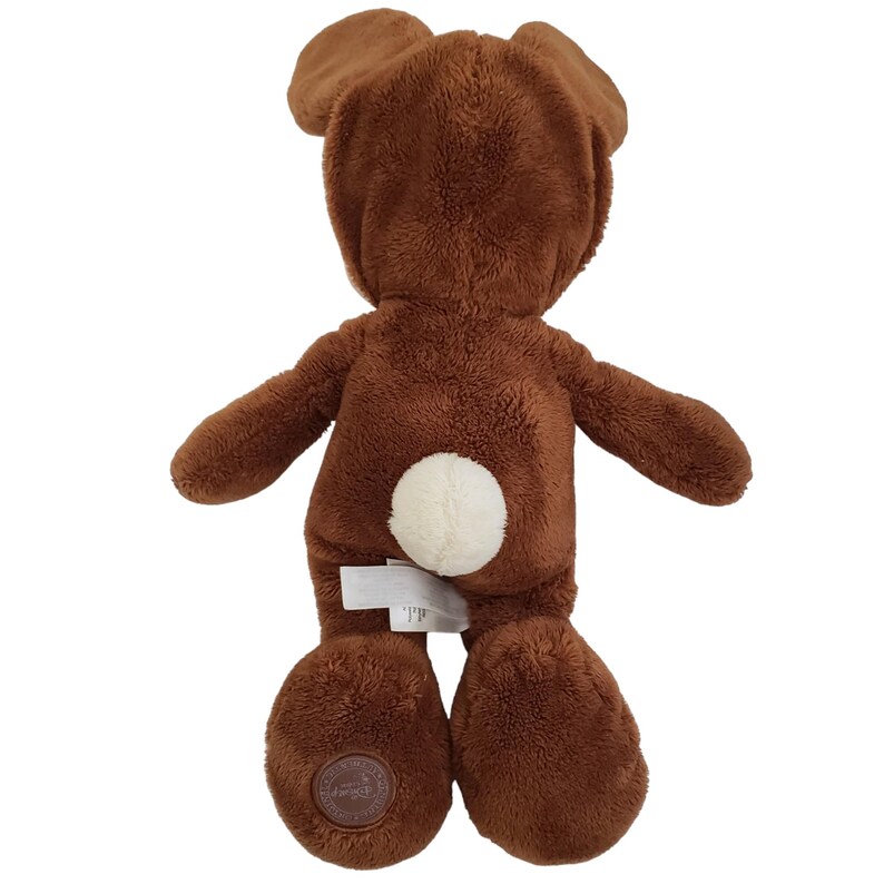 Pode incluir: Um brinquedo de pel&uacute;cia marrom em forma de urso, visto por tr&aacute;s. Possui orelhas grandes, uma cauda branca e fofa e um emblema redondo em um p&eacute;. O brinquedo &eacute; feito de material macio e texturizado.