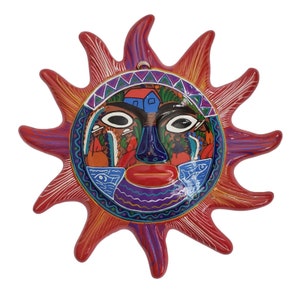 Può includere: Un sole in ceramica rossa con un viso colorato e un design dettagliato. Il viso ha occhi blu, una bocca rossa e un naso viola. Il sole ha un bordo giallo e arancione.