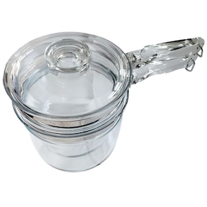 Könnte beinhalten: Ein klarer Glas-Doppelkessel mit Glasdeckel und einem klaren Glasgriff. Der Griff ist mit Metallbeschlägen an der Seite des Topfes befestigt. Der Doppelkocher ist für das Kochen und die Zubereitung von Speisen konzipiert.