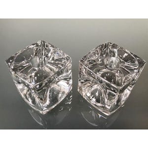 May include: Two clear glass candle holders. Each is a cube shape with a circular indentation in the center for a candle. The glass has a textured, geometric design. The candle holders are displayed on a reflective surface.