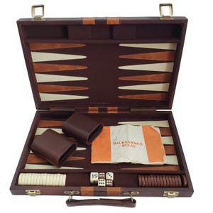 Puede incluir: Juego de backgammon abierto, con exterior de cuero marrón oscuro y superficie de juego beige y blanca. Incluye dados, fichas y un folleto con "Backgammon Rules". El juego tiene un asa y cierres metálicos.