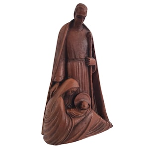 May include: A brown wooden sculpture depicting the Holy Family. The sculpture features a standing figure with a staff, a kneeling woman, and a baby. The figures are draped in flowing robes, and the sculpture has a smooth, carved texture.