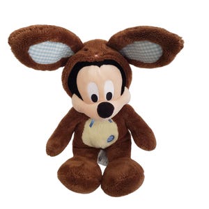 Pode incluir: Um Mickey Mouse de pel&uacute;cia marrom vestido com uma fantasia de urso. A fantasia tem orelhas grandes com um padr&atilde;o xadrez azul e branco por dentro. O rosto do Mickey &eacute; creme com olhos pretos e um nariz preto. Uma fita azul est&aacute; presa.