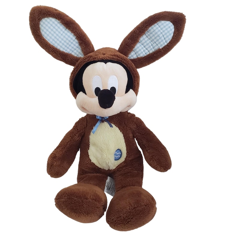 Pode incluir: Um Mickey Mouse de pel&uacute;cia marrom vestido com uma fantasia de coelho. A fantasia apresenta orelhas grandes e flex&iacute;veis com um padr&atilde;o xadrez azul e branco no interior. Mickey tem um rosto bege claro, nariz preto e um la&ccedil;o azul.