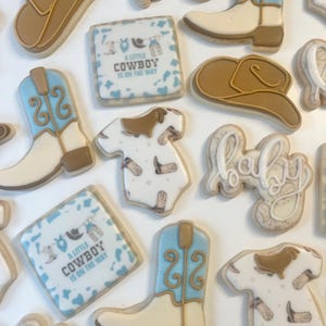 May include: A set of 12 decorated sugar cookies featuring a cowboy theme. The cookies are decorated with blue and brown frosting and include images of cowboy boots, hats, and a baby onesie. The cookies also feature the text "A Little Cowboy Is On The Way".