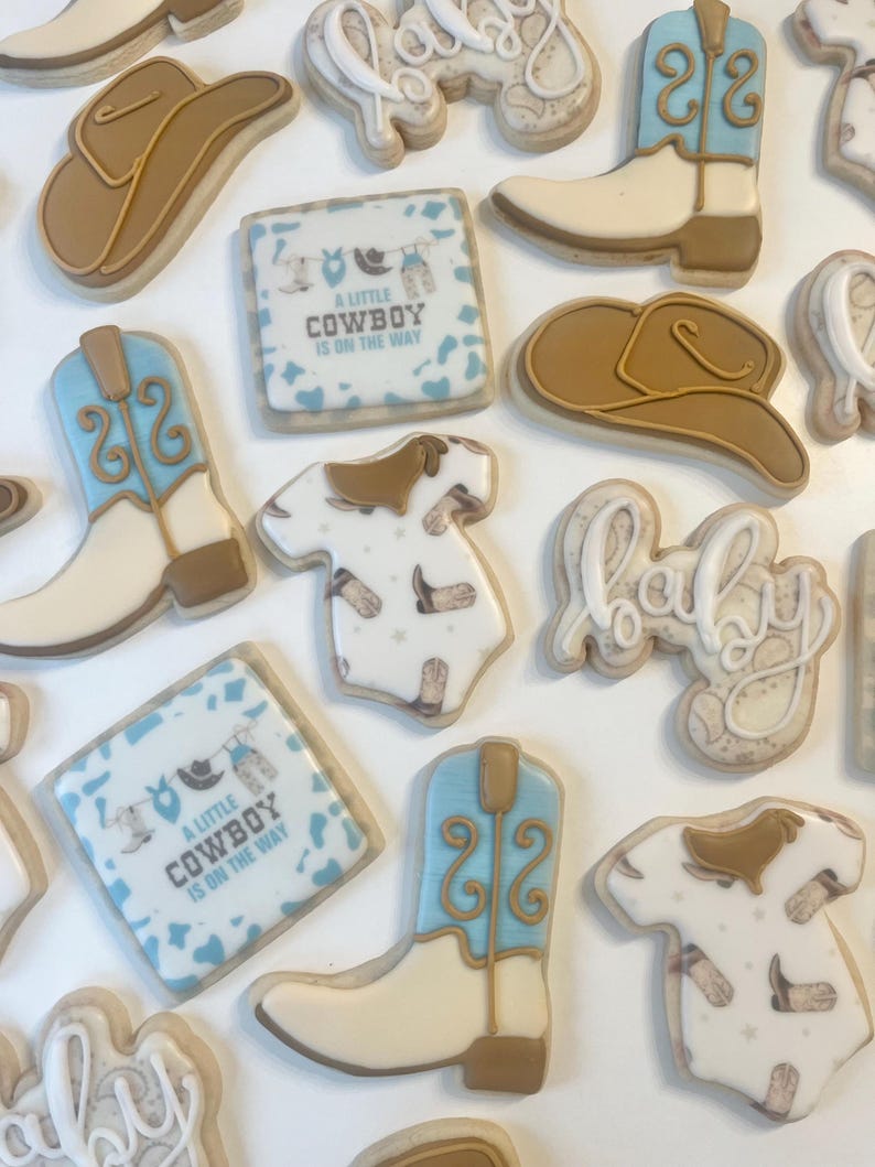 May include: A set of 12 decorated sugar cookies featuring a cowboy theme. The cookies are decorated with blue and brown frosting and include images of cowboy boots, hats, and a baby onesie. The cookies also feature the text "A Little Cowboy Is On The Way".