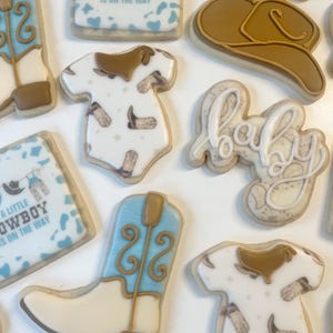 May include: A set of nine decorated sugar cookies featuring a cowboy boot, a baby onesie, a cowboy hat, a baby bib, and the word "baby". The cookies are decorated with blue, brown, and white icing and have a cowboy theme.