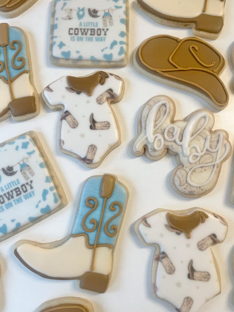 May include: A set of nine decorated sugar cookies featuring a cowboy boot, a baby onesie, a cowboy hat, a baby bib, and the word "baby". The cookies are decorated with blue, brown, and white icing and have a cowboy theme.