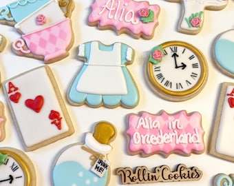May include: Assortment of Alice in Wonderland themed cookies. Includes cookies shaped like clocks, teacups, mushrooms, a dress, and playing cards. Some cookies have the name "Alia" and the phrase "Alia in Onederland" written on them.
