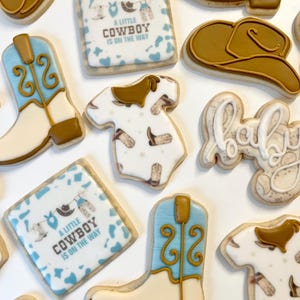 May include: A set of nine decorated sugar cookies with a cowboy theme. The cookies feature a baby onesie, cowboy boots, a cowboy hat, and a sign that says "A little cowboy is on the way".