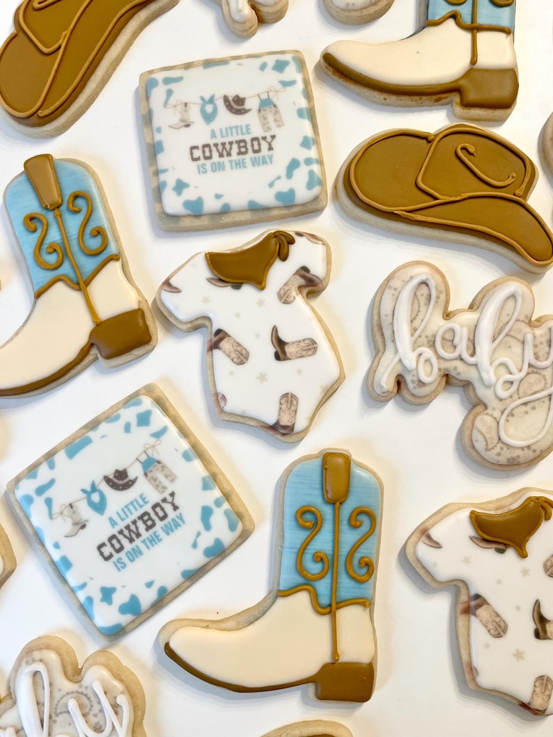 May include: A set of nine decorated sugar cookies with a cowboy theme. The cookies feature a baby onesie, cowboy boots, a cowboy hat, and a sign that says "A little cowboy is on the way".