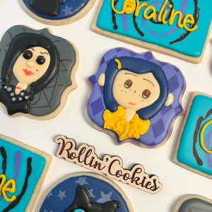 May include: A set of decorated cookies featuring characters from the movie Coraline. The cookies are decorated with blue, purple, and gray icing and feature images of Coraline, the Other Mother, and the black cat. The cookies are arranged on a white surface with a wooden sign that reads "Rollin' Cookies".