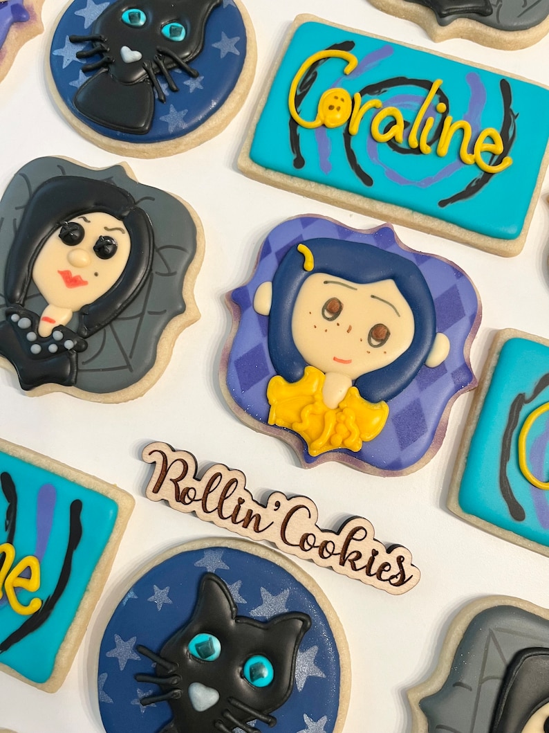 May include: A set of decorated cookies featuring characters from the movie Coraline. The cookies are decorated with blue, purple, and gray icing and feature images of Coraline, the Other Mother, and the black cat. The cookies are arranged on a white surface with a wooden sign that reads "Rollin' Cookies".