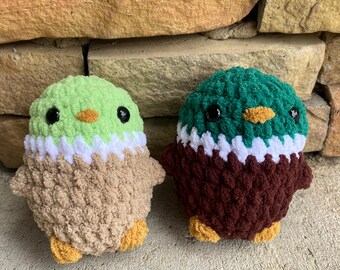 May include: Two handmade crochet ducklings. One is light green, white, and tan, the other is dark green, white, and brown. Both have black eyes and orange beaks. The ducklings are sitting on a concrete surface in front of a stone wall.