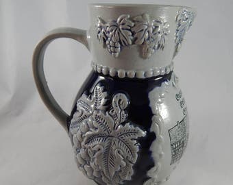 Puede incluir: Una jarra de cerámica azul y blanca con asa y diseño de vid. La jarra tiene un esmalte azul y detalles en relieve blancos. El texto "Oberammergau" es visible en el lateral.