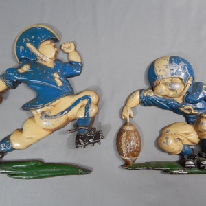 Può includere: Due pezzi di arredamento da parete in metallo vintage di giocatori di football americano. Un giocatore corre con la palla e l'altro è in posizione difensiva. Entrambi i giocatori indossano uniformi blu e bianche con caschi.