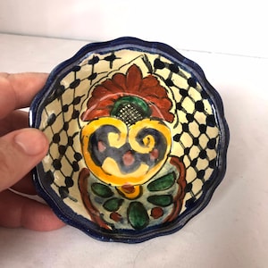 Op de afbeelding: Een kleine, ronde, keramische kom met een gegolfde rand. De kom is versierd met een kleurrijk bloemenontwerp in tinten blauw, groen, geel, rood en wit. Het ontwerp heeft een centrale bloem met een gele kern en een witte achtergrond.