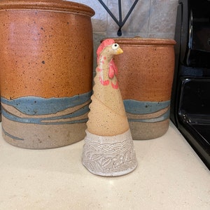 May include: A ceramic chicken figurine with a white base and a pink and brown body. The figurine is standing on a countertop with other ceramic items in the background.