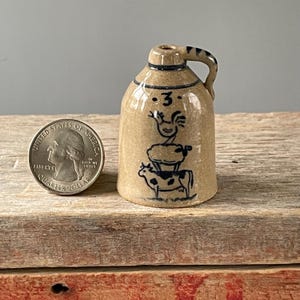 May include: A small, tan ceramic jug with a handle and a blue stripe around the top. The jug is decorated with a blue illustration of a rooster, a stack of hay, and a cow. The number "3" is above the rooster. A quarter is next to the jug.