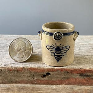 May include: A small, tan ceramic jar with a blue bee design and blue accents. The jar has two small handles and a decorative blue band around the top. A quarter is next to the jar for size comparison.