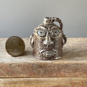 May include: A small, brown ceramic jug with a face sculpted on the front. The jug has a handle and a spout. The face has large eyes, a prominent nose, and a wide, toothy grin. A quarter is next to the jug for size comparison.