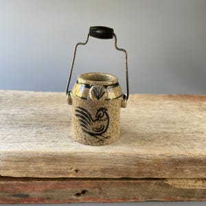 May include: A small ceramic milk can with a black rooster design and a black metal handle. The can is sitting on a wooden surface.