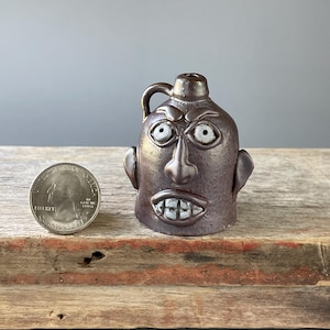 May include: A brown ceramic jug with a face sculpted on the front. The jug has a handle and a small opening at the top. The face has large, white eyes, a prominent nose, and a toothy grin. A quarter is next to the jug for size comparison.