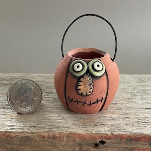 May include: A small, round, terracotta-colored ceramic pumpkin bucket with a black wire handle. The pumpkin features two pale green eyes, a textured tan nose, and a stitched mouth design. A US quarter coin is placed beside it for scale.