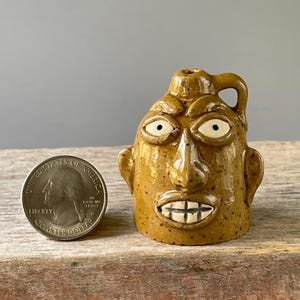 May include: A small, mustard-yellow ceramic face jug with a speckled glaze, featuring wide white eyes, a prominent nose, and bared teeth. The jug is positioned next to a United States quarter coin for scale, highlighting its miniature size.