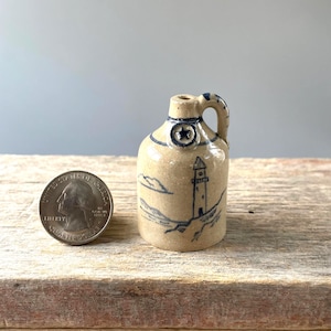 May include: A small, tan ceramic jug with a blue lighthouse design. The jug has a handle and a star emblem. A U.S. quarter coin is next to the jug for size comparison. The jug is on a wooden surface.