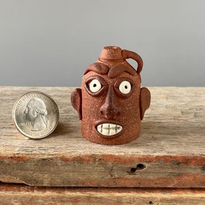 May include: A small, brown ceramic jug with a face sculpted on the front. The face has white eyes, a brown nose, and a white mouth with teeth. The jug has a handle on the side.