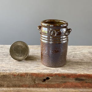 May include: A small, handcrafted ceramic vase with a dark brown glaze. The vase features two small handles and a decorative emblem. The vase is approximately 2.5 inches tall and sits on a wooden surface.