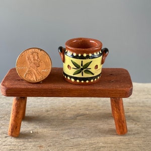 May include: A small, hand-painted ceramic pot with a yellow, green, and black design. The pot is sitting on a wooden stool.