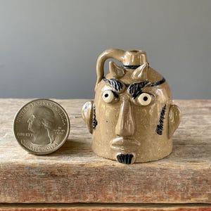 May include: A tan ceramic jug with a face sculpted on the front. The jug has a handle, two small horns, and black accents around the eyes and on the eyebrows. The jug is next to a quarter.