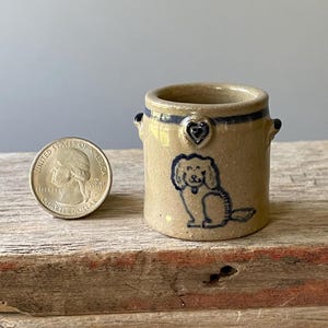 May include: A small tan ceramic crock with two side handles, featuring a dark blue hand-painted dog illustration and a heart motif near the rim. A United States quarter coin is placed next to the crock for scale, showing its miniature size.