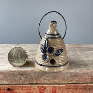 May include: A ceramic beehive-shaped birdhouse with a black metal handle. The birdhouse is tan with blue floral and leaf accents. A U.S. quarter is next to the birdhouse for size comparison.