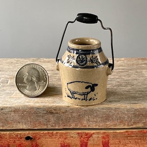 May include: A small, tan ceramic bucket with a black handle and a black handle grip. The bucket has blue painted details, including a sheep illustration and floral designs. A U.S. quarter is next to the bucket for size comparison.