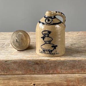 May include: A small, tan ceramic jug with a handle and a dark blue star design. The jug features a hand-painted design of three sheep. A U.S. quarter is next to the jug for size comparison. The jug is on a wooden surface.