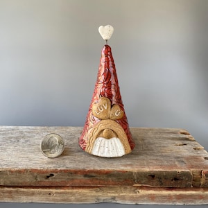 May include: A ceramic gnome figurine with a red and brown hat, a white beard, and two hearts for eyes that say "LUV U". The gnome is wearing a white heart on top of its hat.