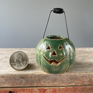 May include: A green ceramic pumpkin-shaped container with a carved jack-o'-lantern face. It has a black handle and a metal wire. A US quarter is next to the pumpkin. The pumpkin is on a wooden surface.